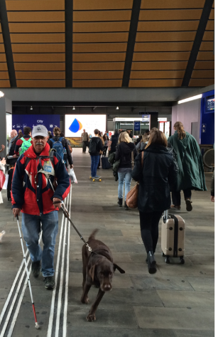 Eine sehbeeinträchtigte Person mit Blindenhund unterwegs auf der Passerelle am Bahnhof SBB Basel